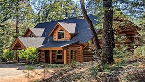 A log cabin with a steeply pitched roof, a front porch, and a dirt driveway.