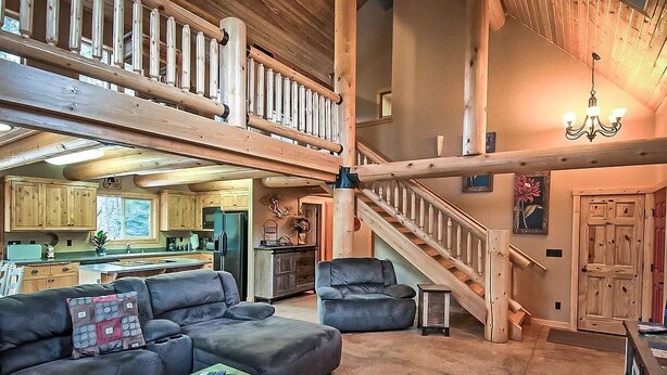 A spacious living area with a high ceiling, wooden beams, a staircase, and a kitchen in the background.