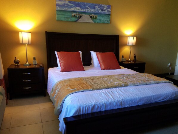 A hotel room with a large bed, two bedside tables with lamps, a nightstand, a wooden headboard, and a painting of a pier over water on the wall.