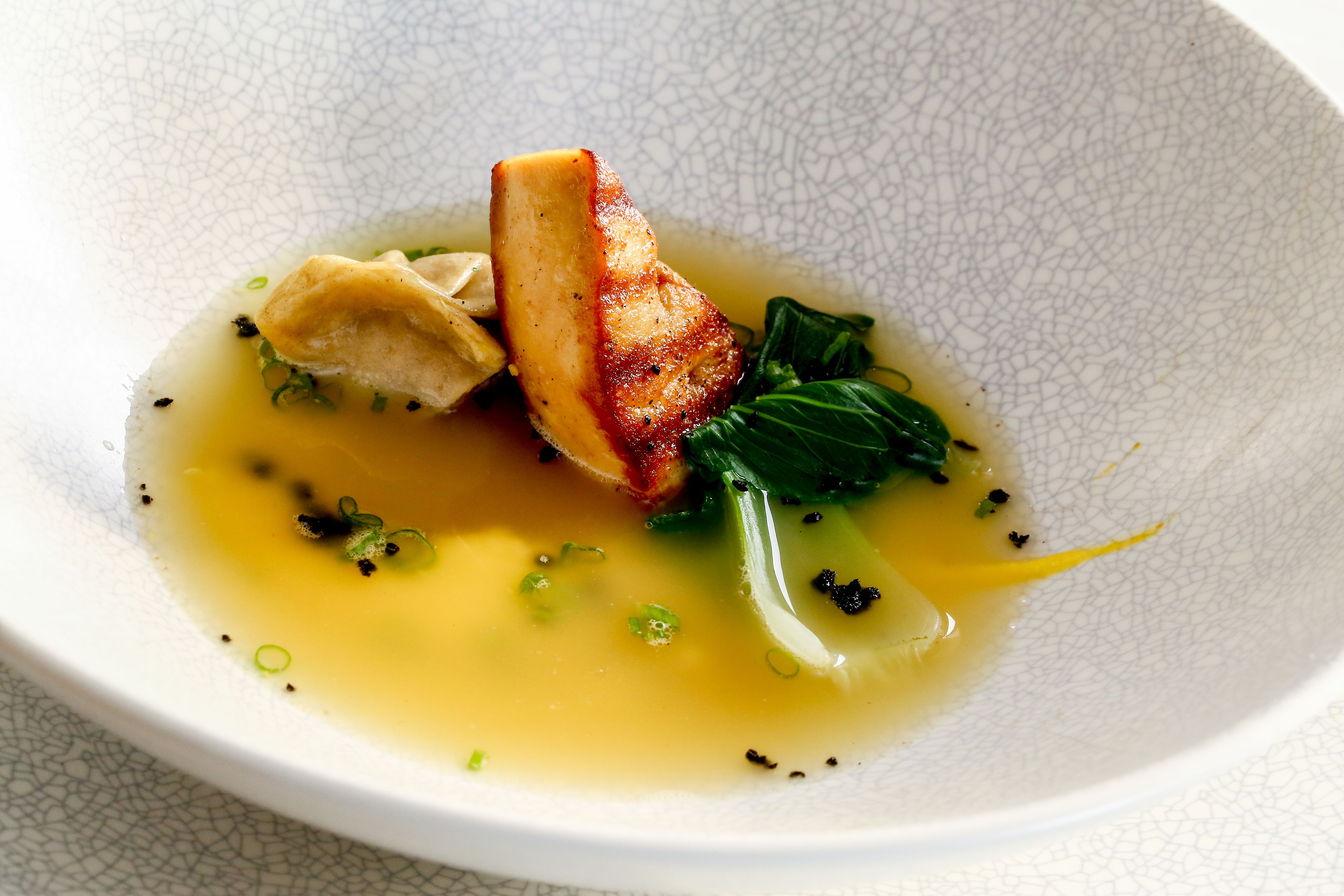 A white bowl with a textured surface containing a dish with a piece of meat, greens, and a clear broth.