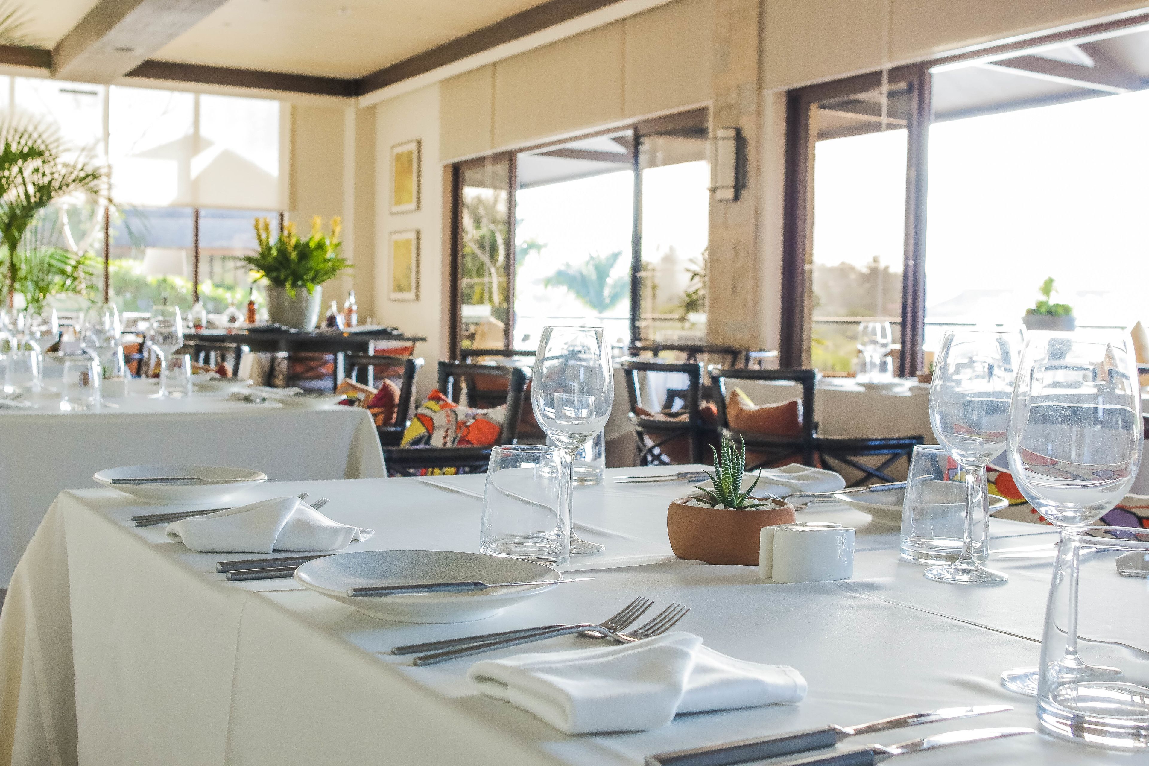 A dining table set with white tablecloths, plates, cutlery, and wine glasses, with potted plants and a view of the outdoors.