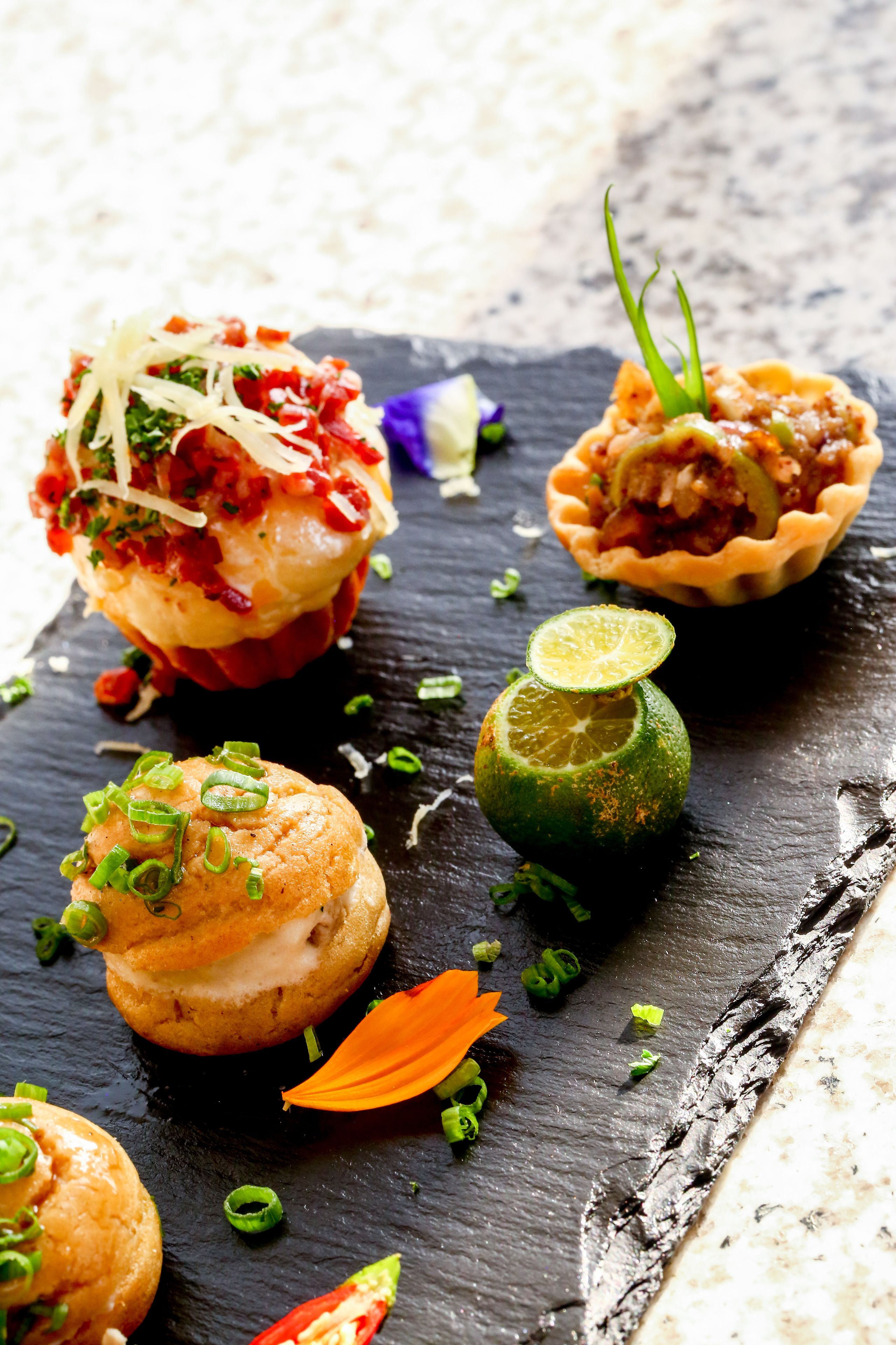 A variety of food items on a slate serving board, including a pastry with red filling, a tart, and a lime garnish.