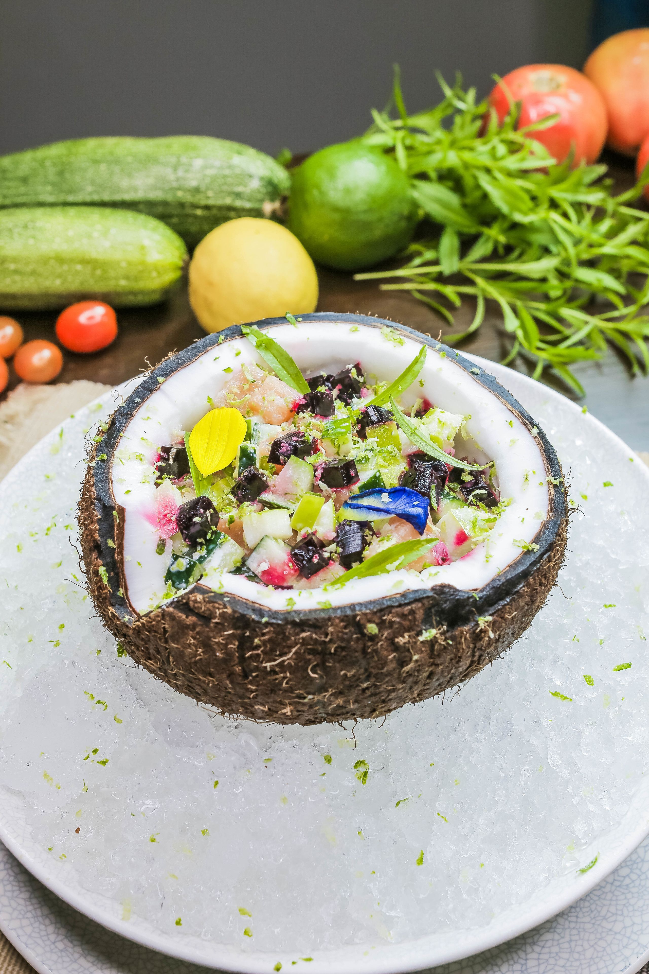 A salad in a coconut shell on a plate with ice, surrounded by vegetables and herbs.