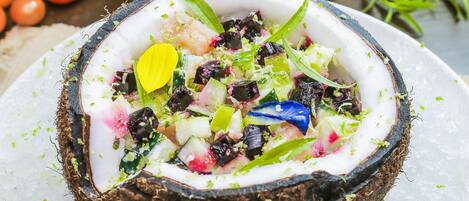 A salad in a coconut shell on a plate with ice, surrounded by vegetables and herbs.
