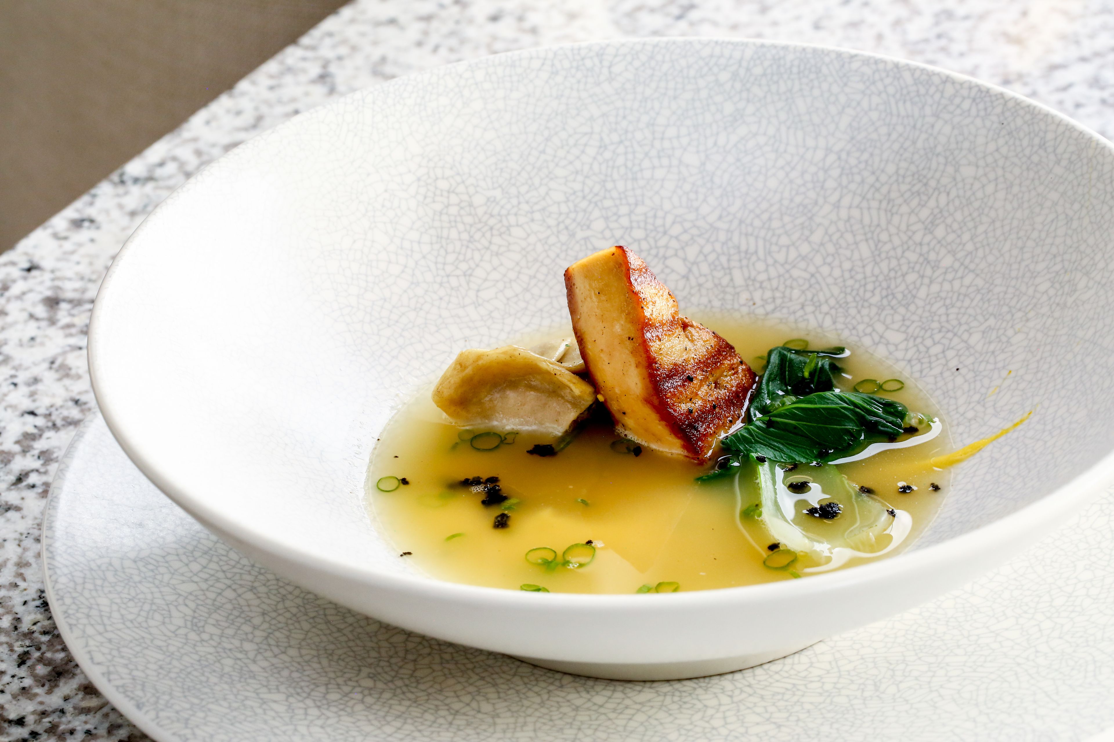 A bowl of soup with a piece of meat and greens on a textured white plate.