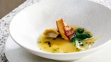 A bowl of soup with a piece of meat and greens on a textured white plate.