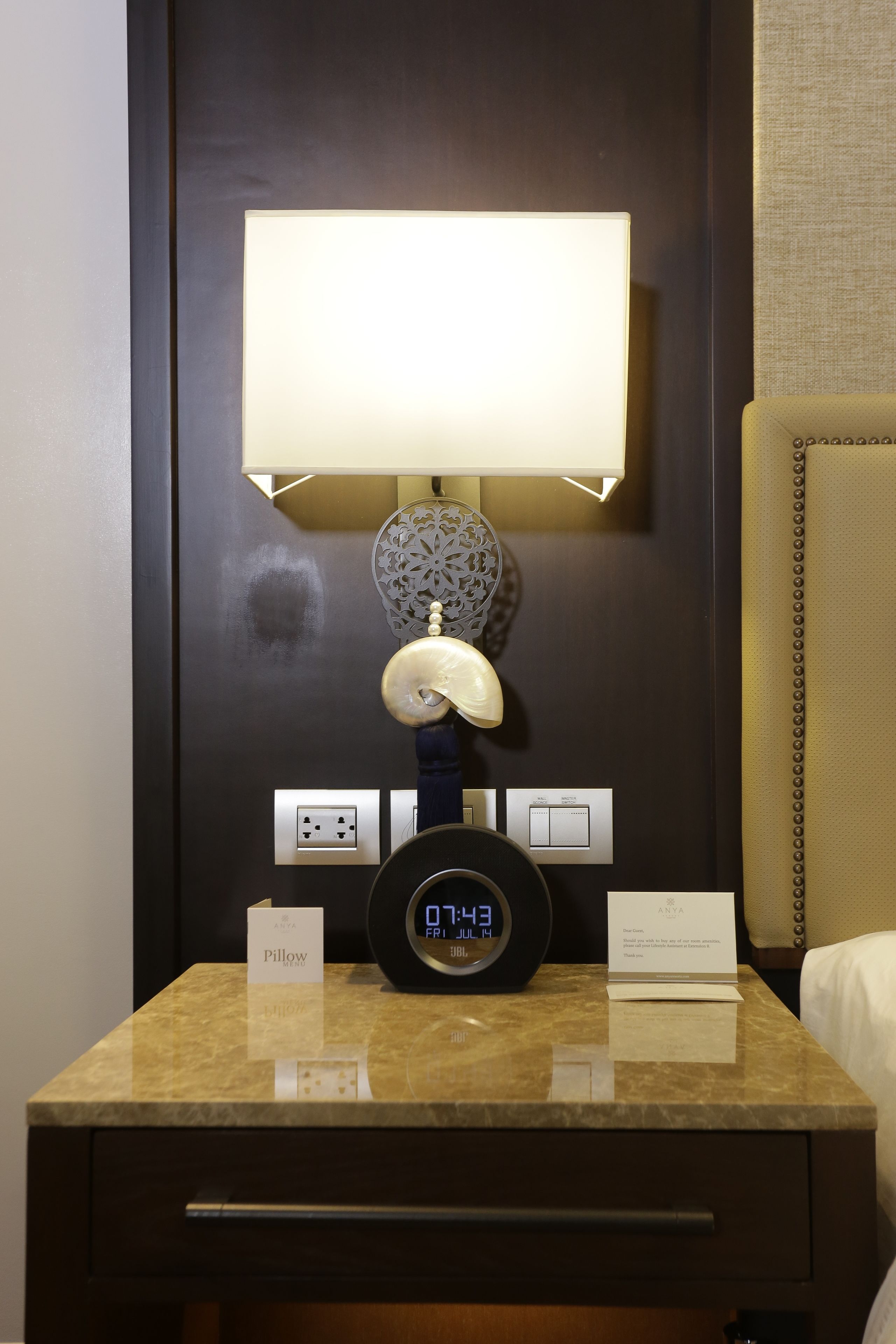 A hotel room with a bedside table featuring a digital clock, a lamp, and a pillow.