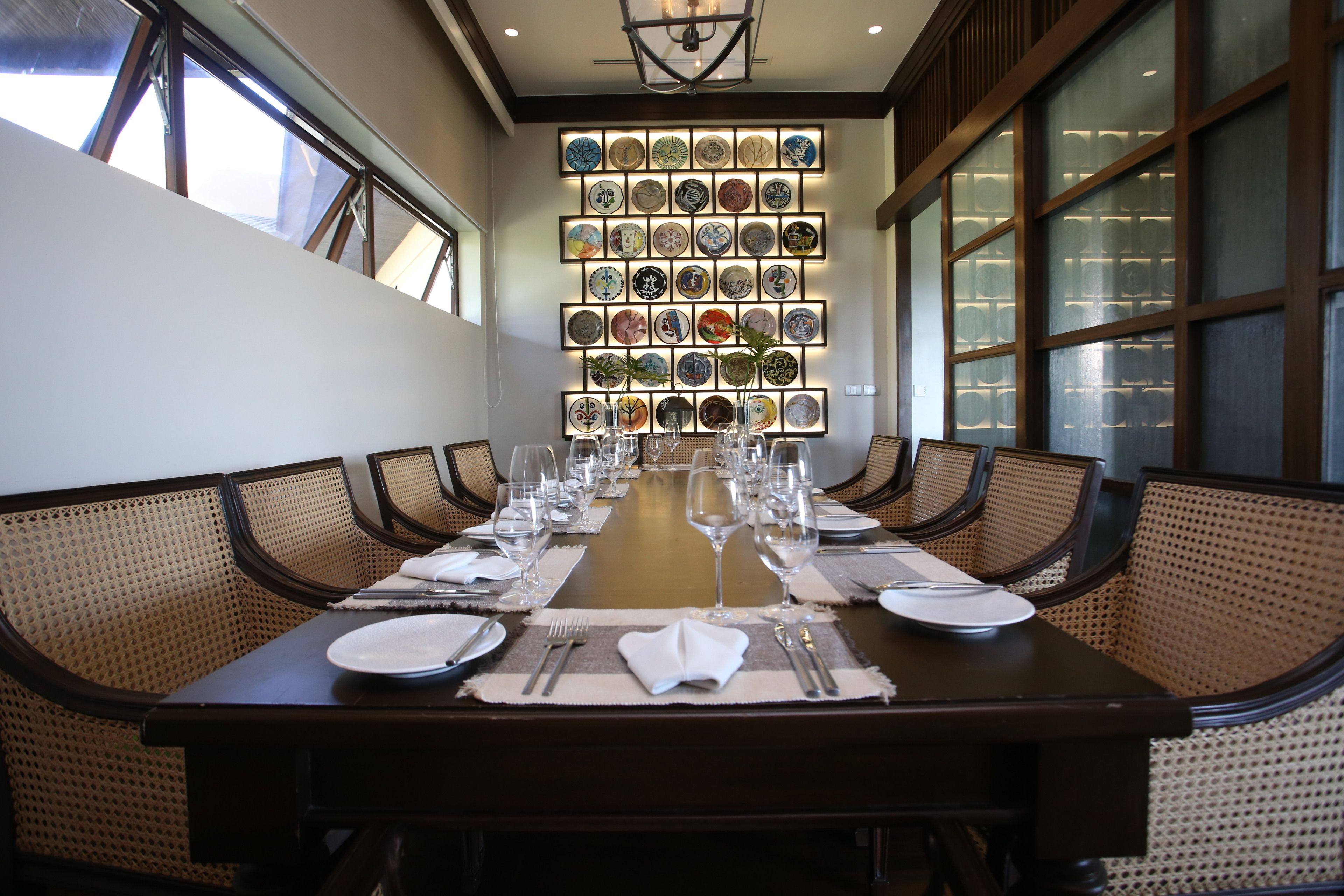 A dining room with a long table set for a meal, surrounded by wicker chairs, and a wall of decorative plates.