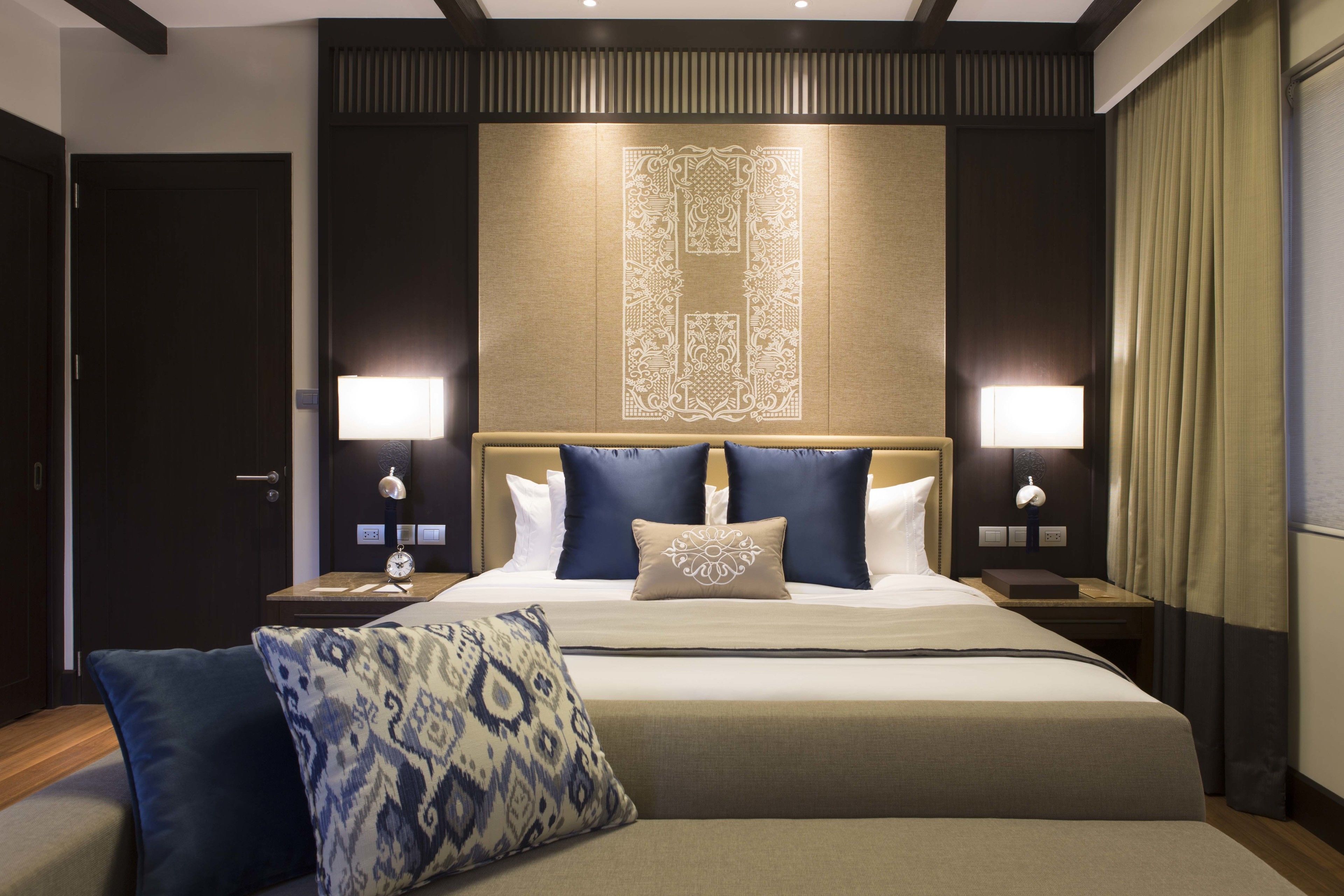 A hotel room with a large bed, two bedside tables, a nightstand, a wall-mounted lamp, and a patterned throw pillow.