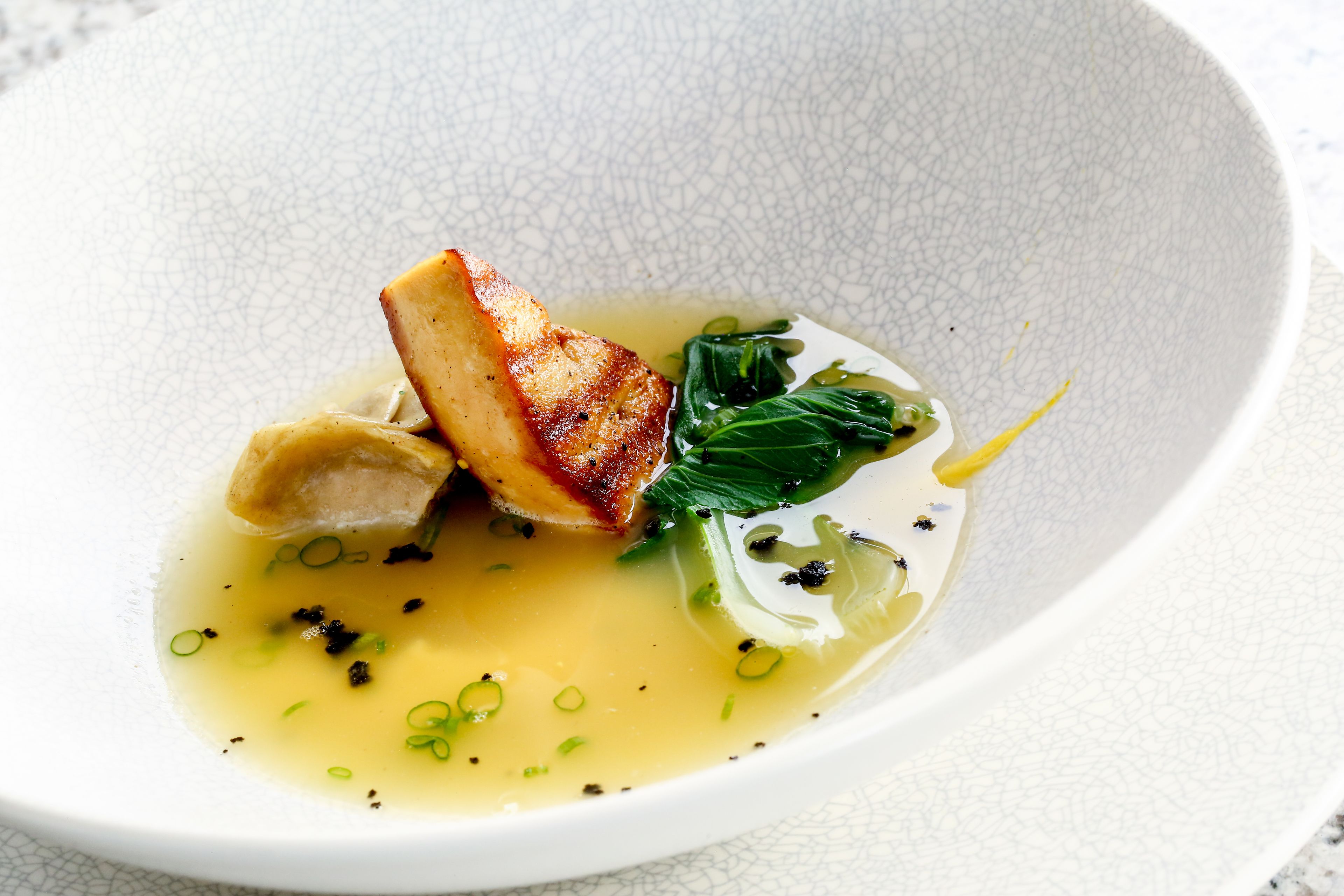 A white ceramic plate with a piece of meat, green vegetables, and a clear broth.