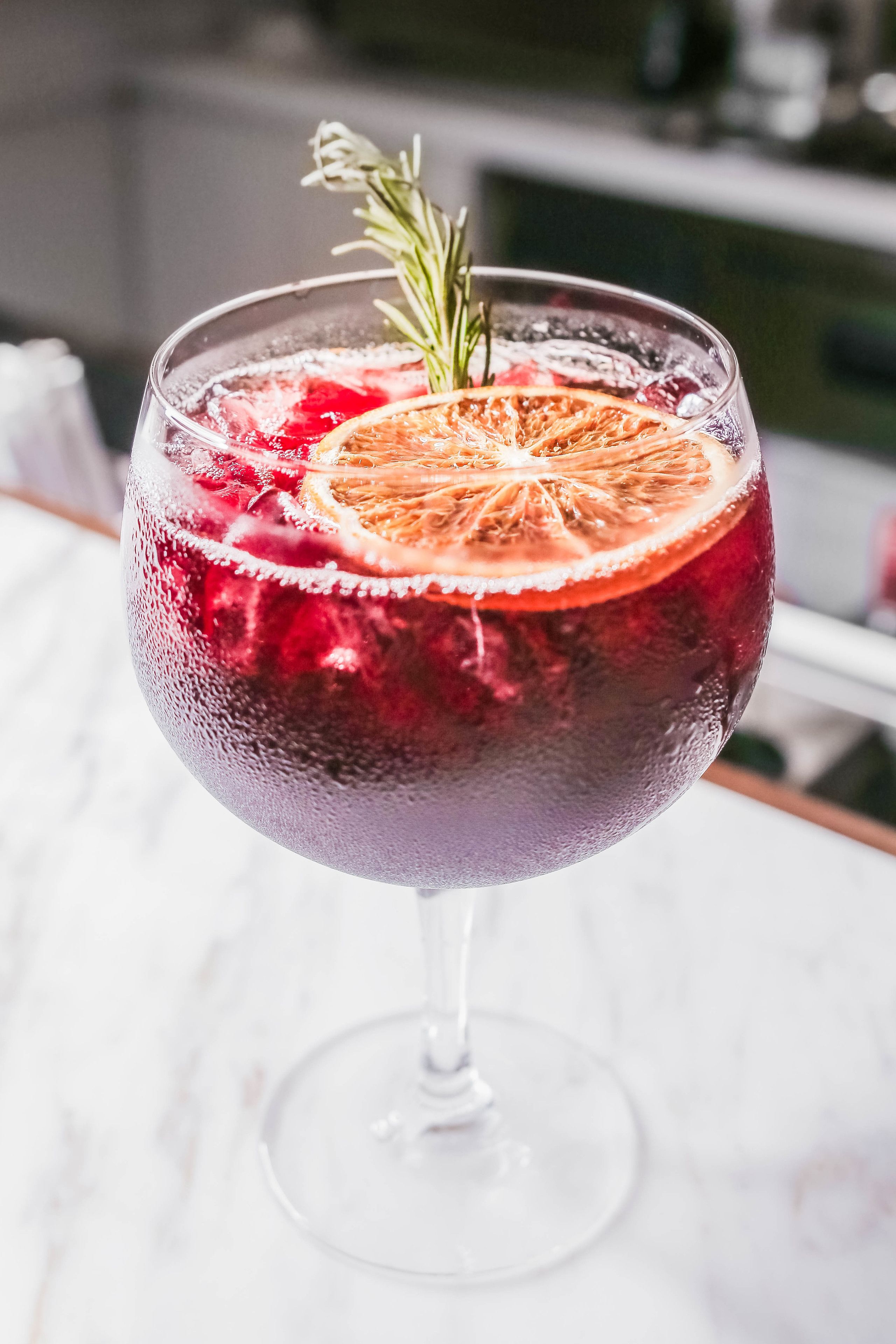 Un verre d’une boisson rouge, servie avec des glaçons, et décorée d’une tranche d’orange et d’une branche de romarin.