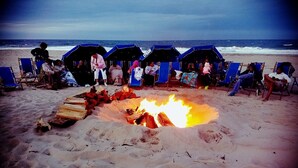 Escena de playa con una hoguera rodeada de sillas y tiendas de playa azules.
