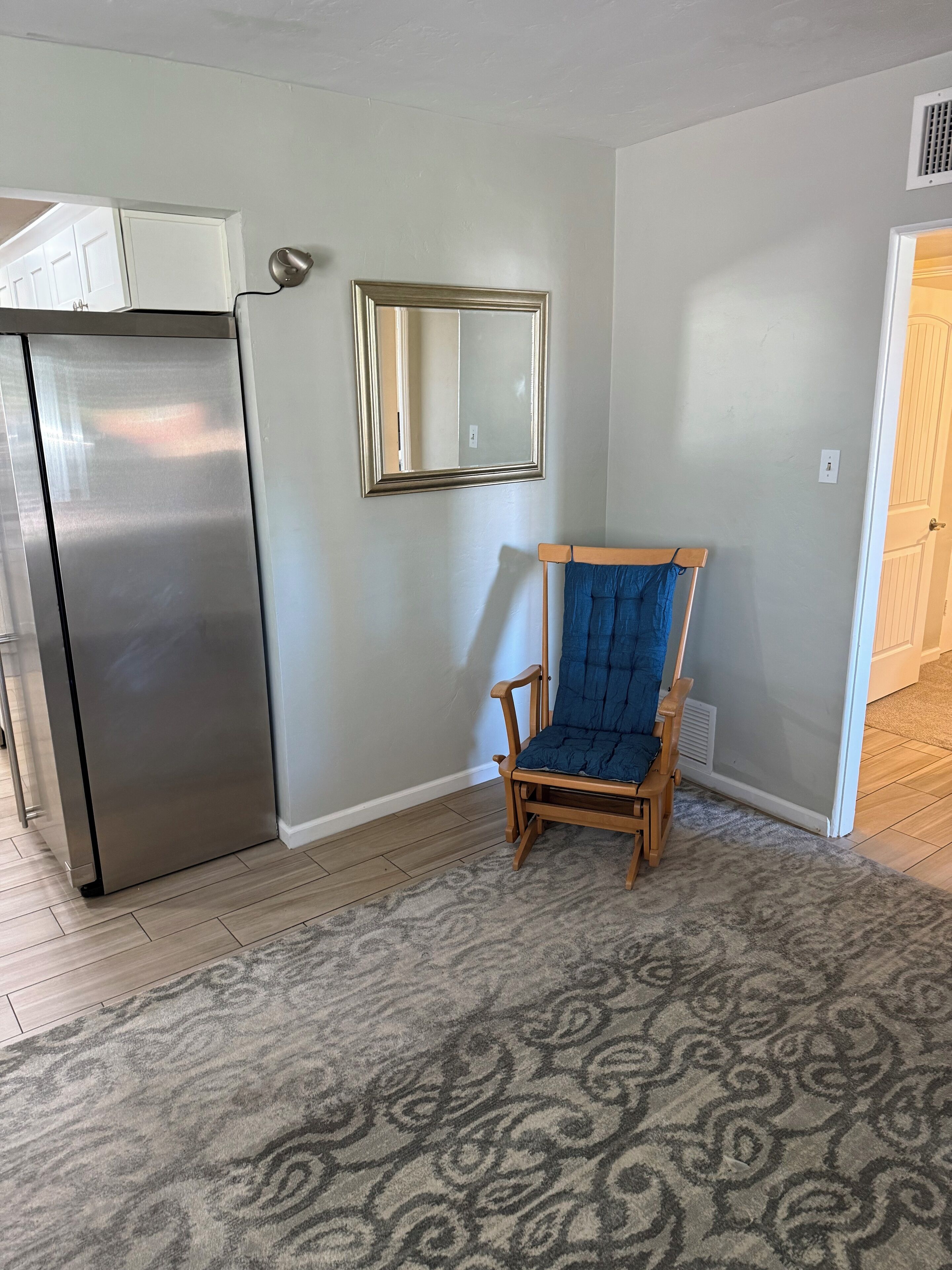 A kitchen with a stainless steel refrigerator, a wooden chair with a blue cushion, and a patterned rug on the floor.