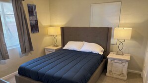A neatly made bed with a dark blue comforter and white pillows, flanked by two bedside tables with lamps, a remote control on a side table, and a window with curtains. - Beautifully Charming Redbrick Home (Phoenix)
