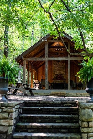 A rustic wooden cabin with a stone fireplace, surrounded by trees and a stone staircase leading to the entrance.
