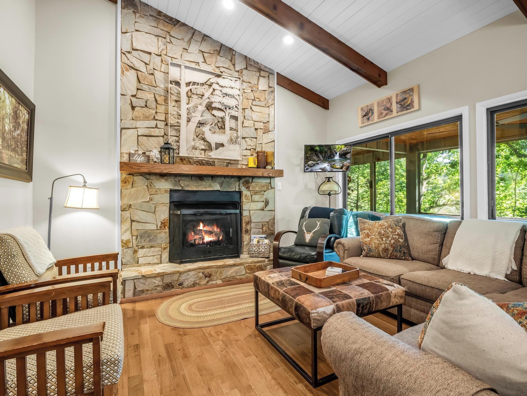 A cozy living room with a stone fireplace, comfortable seating, and a large window offering a view of the outdoors.