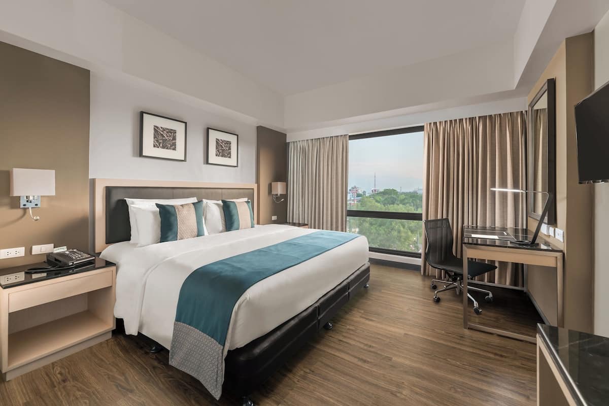 Ein modernes Hotelzimmer mit einem großen Bett, einem Schreibtisch mit einem Laptop, einem Fernseher und Blick auf Grünflächen.