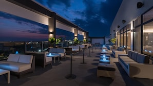 An outdoor seating area with multiple couches and tables, illuminated by ambient lighting, set against a backdrop of a twilight sky.