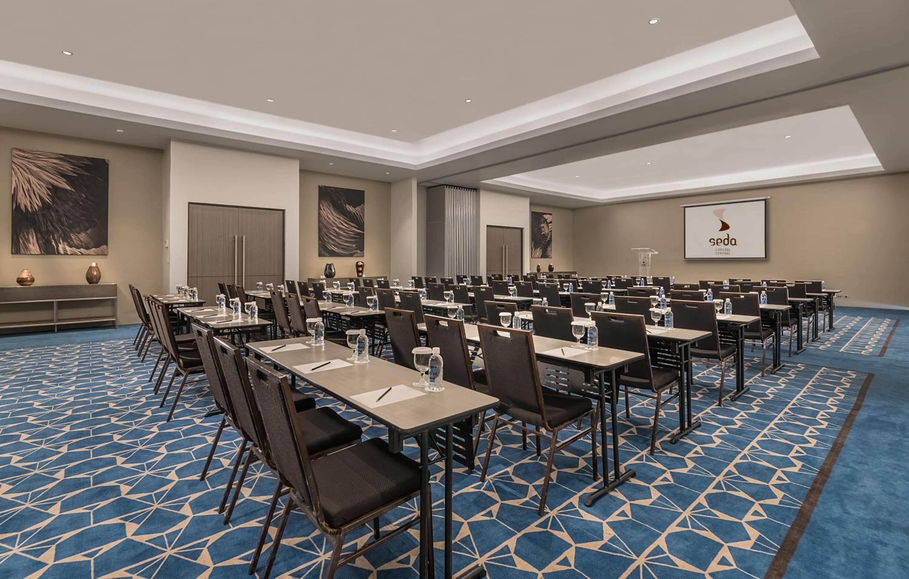 A conference room with rows of tables and chairs, a podium, and a screen displaying the logo 'seda'.