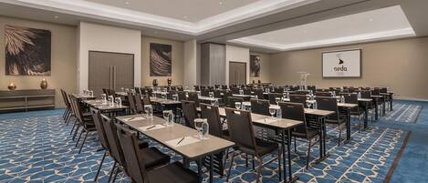 A conference room with rows of tables and chairs, a podium, and a screen displaying the logo 'seda'.