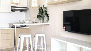 A modern kitchen with white cabinetry, a gas stove, two white stools, a hanging light fixture, and a mounted flat-screen TV. - Pinnacle PJ Penguin Homes (Petaling Jaya)