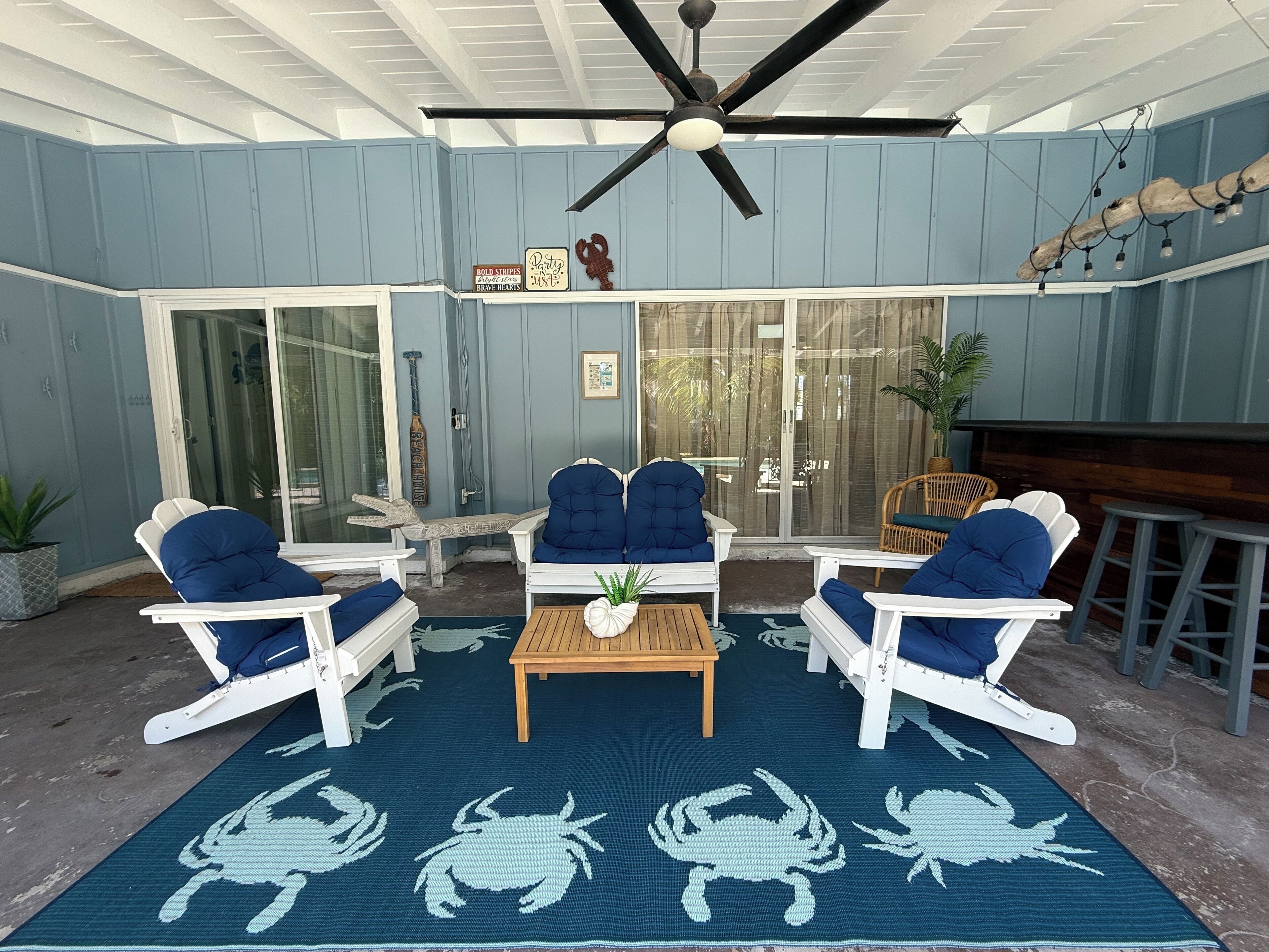 A covered patio area with white furniture, a ceiling fan, and a blue crab-patterned rug.
