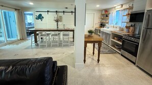 Eine moderne Küche mit Edelstahlgeräten, ein Essbereich mit weißen Stühlen und ein Wohnzimmer mit einem schwarzen Ledersofa.