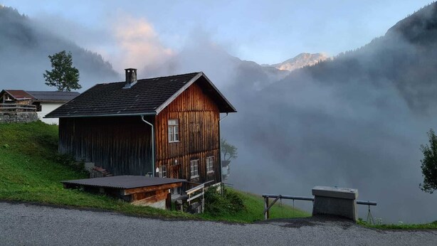 A rustic wooden cabin with a sloped roof, situated on a grassy hill with a view of mist-covered mountains in the background. - Apartment for 2 - 3 people (Wängle)