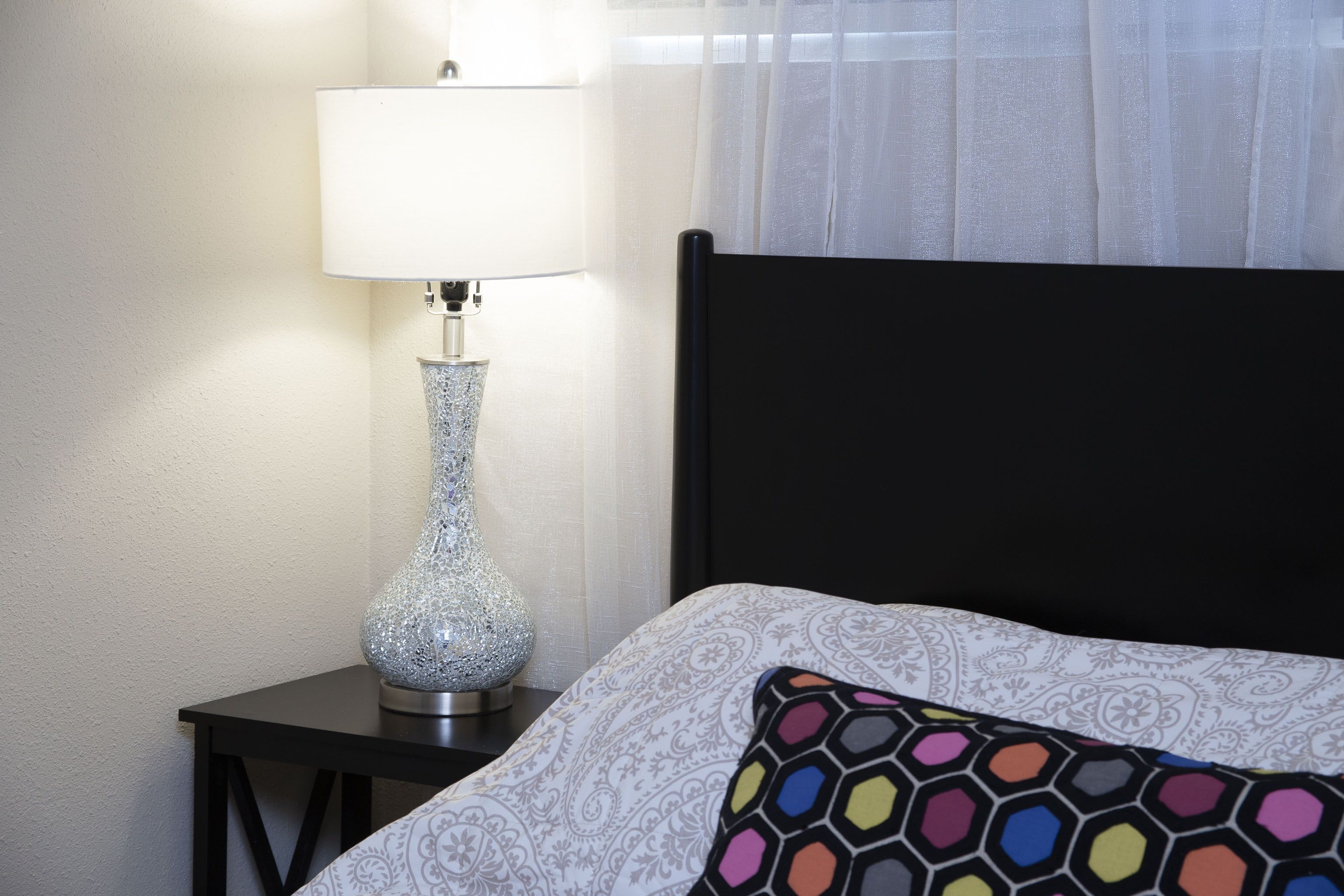 A bed with a patterned pillow, a decorative lamp on a bedside table, and a sheer curtain in the background.