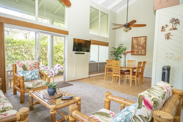 A spacious living area with a dining set, a ceiling fan, and a large window overlooking a garden.