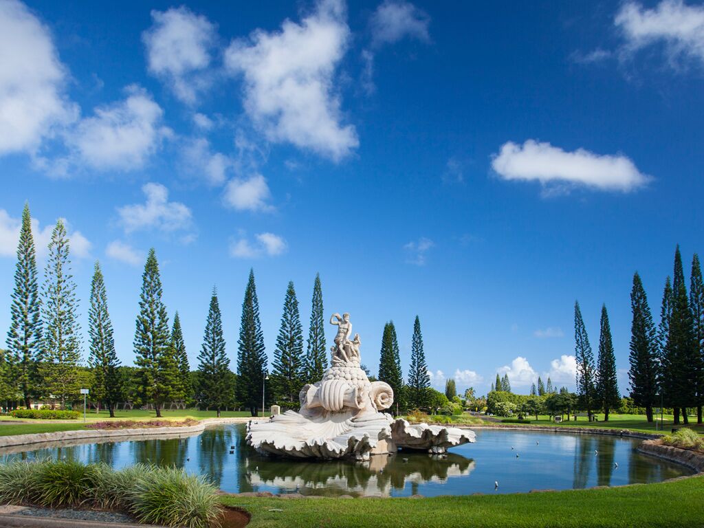 Une fontaine ornée d’une statue au centre, entourée d’un bassin et de grands arbres.