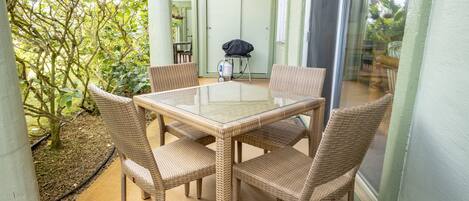 Een tafel met glazen blad en rieten stoelen in een overdekte buitenruimte met uitzicht op groen.