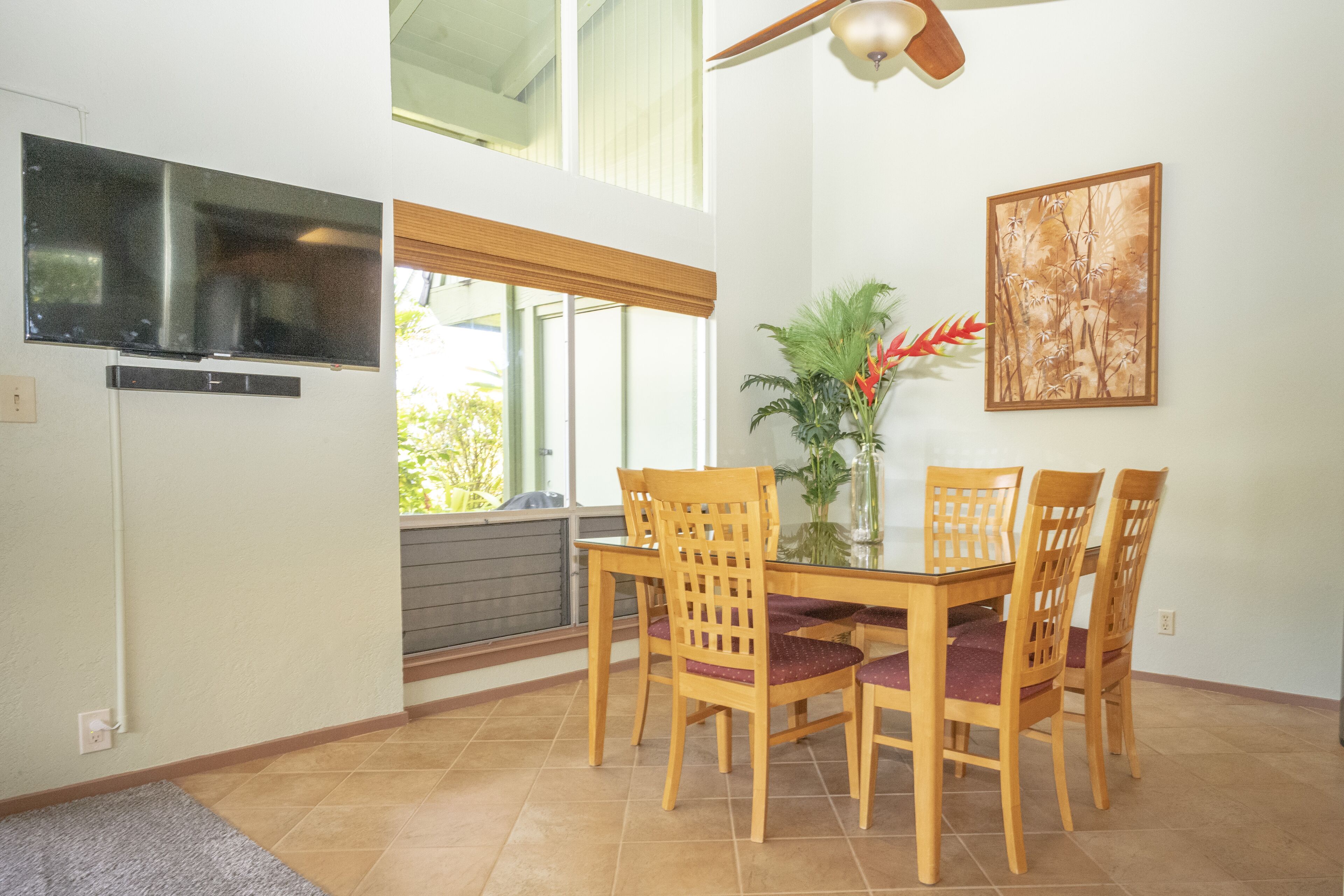 Un comedor con mesa y sillas, un televisor montado en la pared y una ventana con persianas.