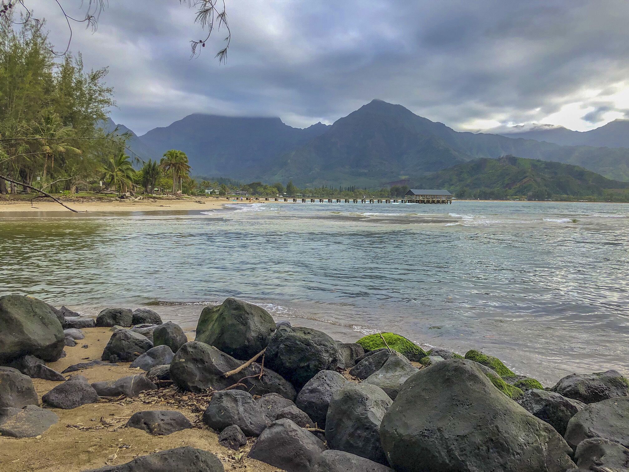A beach with large rocks in the foreground, a pier extending into the water, and mountains in the background.