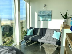 Una sala de estar moderna con un sofá gris, vista a la playa y un cuadro en la pared.