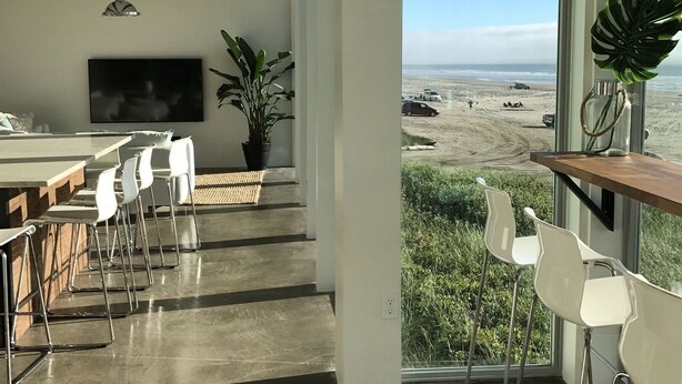 Interior moderno com uma janela grande com vista para uma praia, área de refeições com cadeiras brancas e uma planta em vaso.