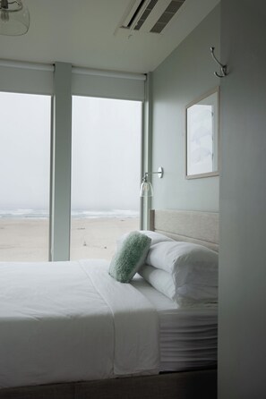 A bedroom with a bed, a window with a view of the beach, and a wall-mounted light fixture. - Cleanline Beach House: Modern Oceanfront Luxury (Cloverdale)
