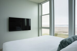 Une chambre à coucher avec un lit, un téléviseur fixé au mur et une grande fenêtre donnant sur la plage.