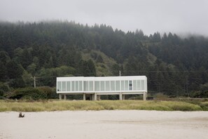 Un bâtiment moderne doté d’un toit plat, de grandes fenêtres et de fondations en béton, situé dans une zone herbeuse, avec une colline boisée en arrière-plan.