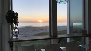 Quarto com vista para o pôr do sol sobre o oceano, um vaso de vidro com uma planta e uma mesa com cadeiras.