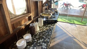 A buffet setup with a variety of dishes and utensils on a stone-tiled counter, surrounded by wooden walls and a large window.