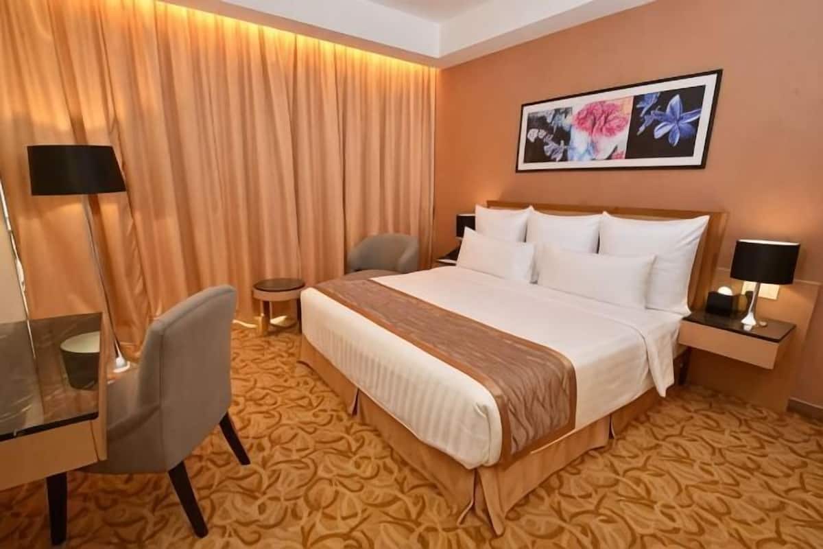 A hotel room with a large bed, a desk with a chair, a small table, and a nightstand with a lamp.
