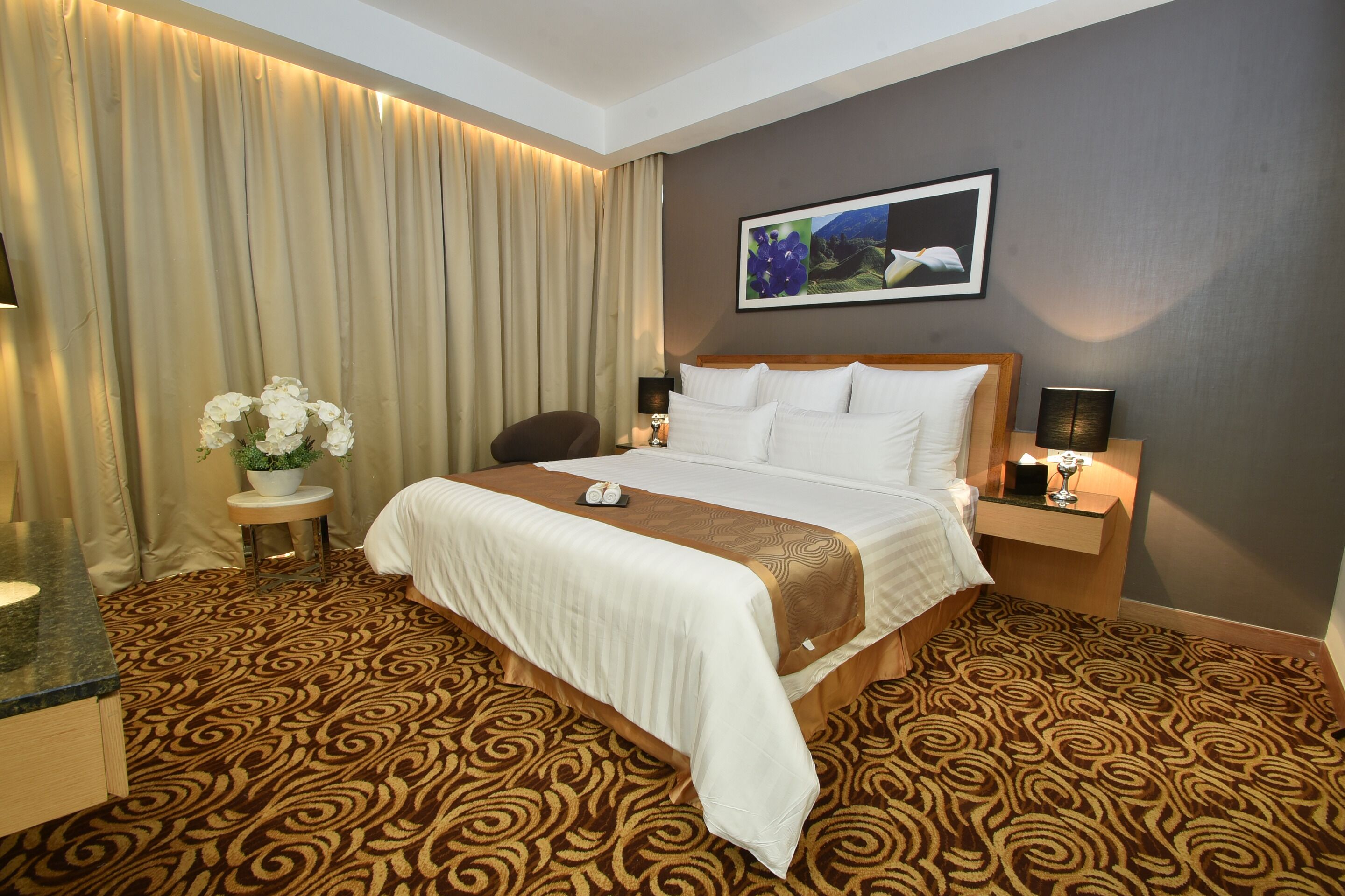 A hotel room with a large bed, a bedside table with a lamp, a small round table with a vase of flowers, and a wall-mounted picture.