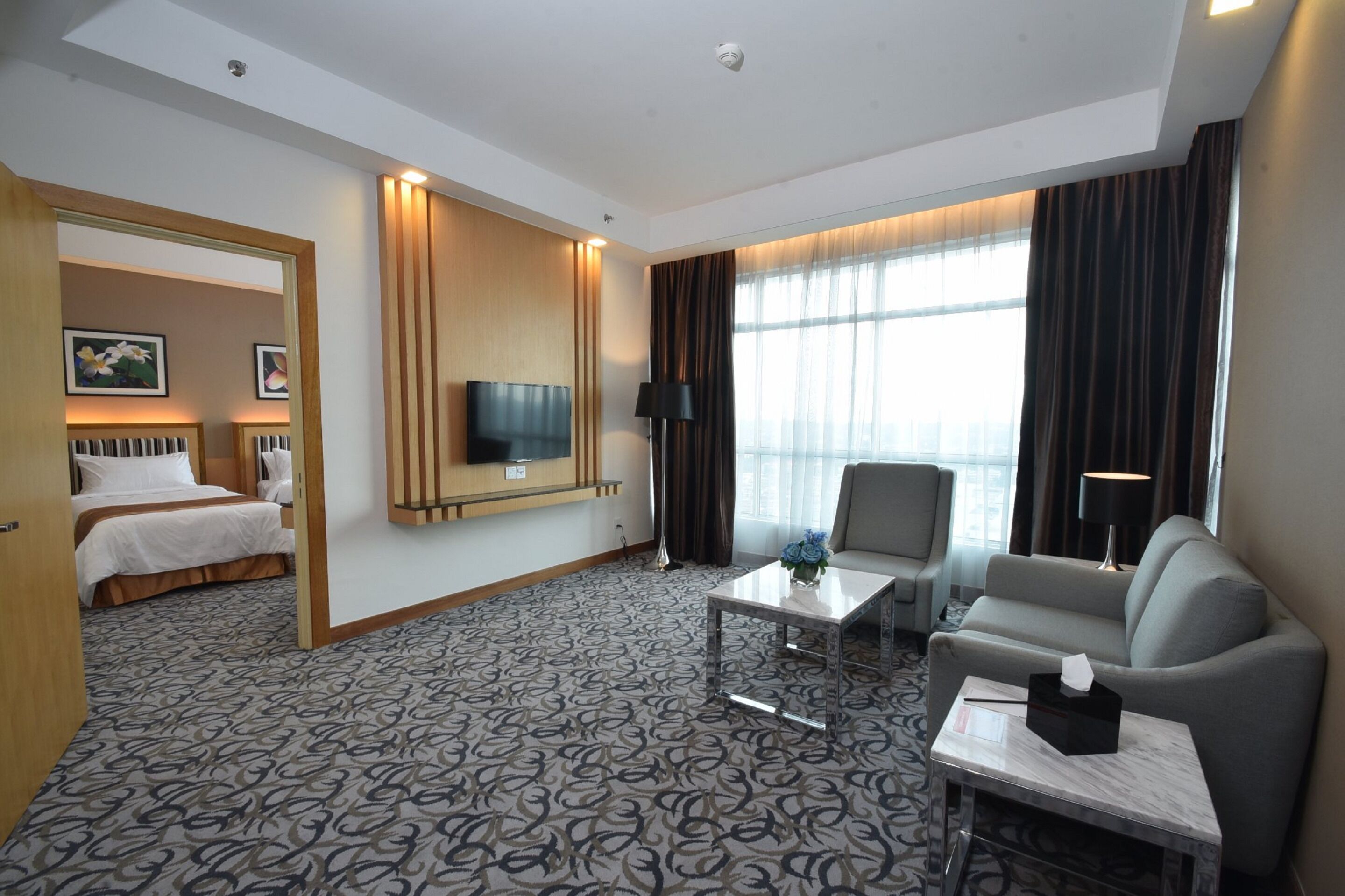 Habitación de hotel con cama, sofá, televisor, mesa de centro y ventana con cortinas.