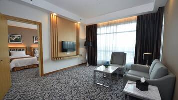 Habitación de hotel con cama, sofá, televisor, mesa de centro y ventana con cortinas.