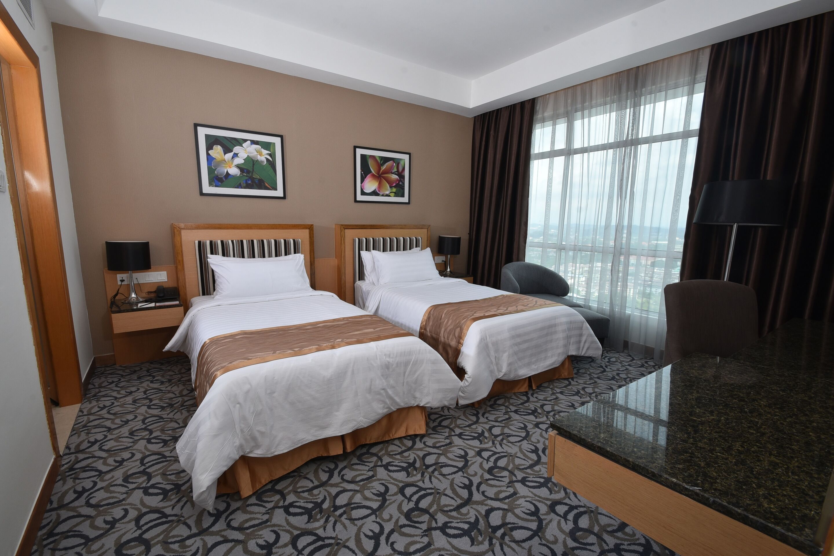 A hotel room with two beds, a desk, a chair, a large window with curtains, and two framed floral paintings on the wall.