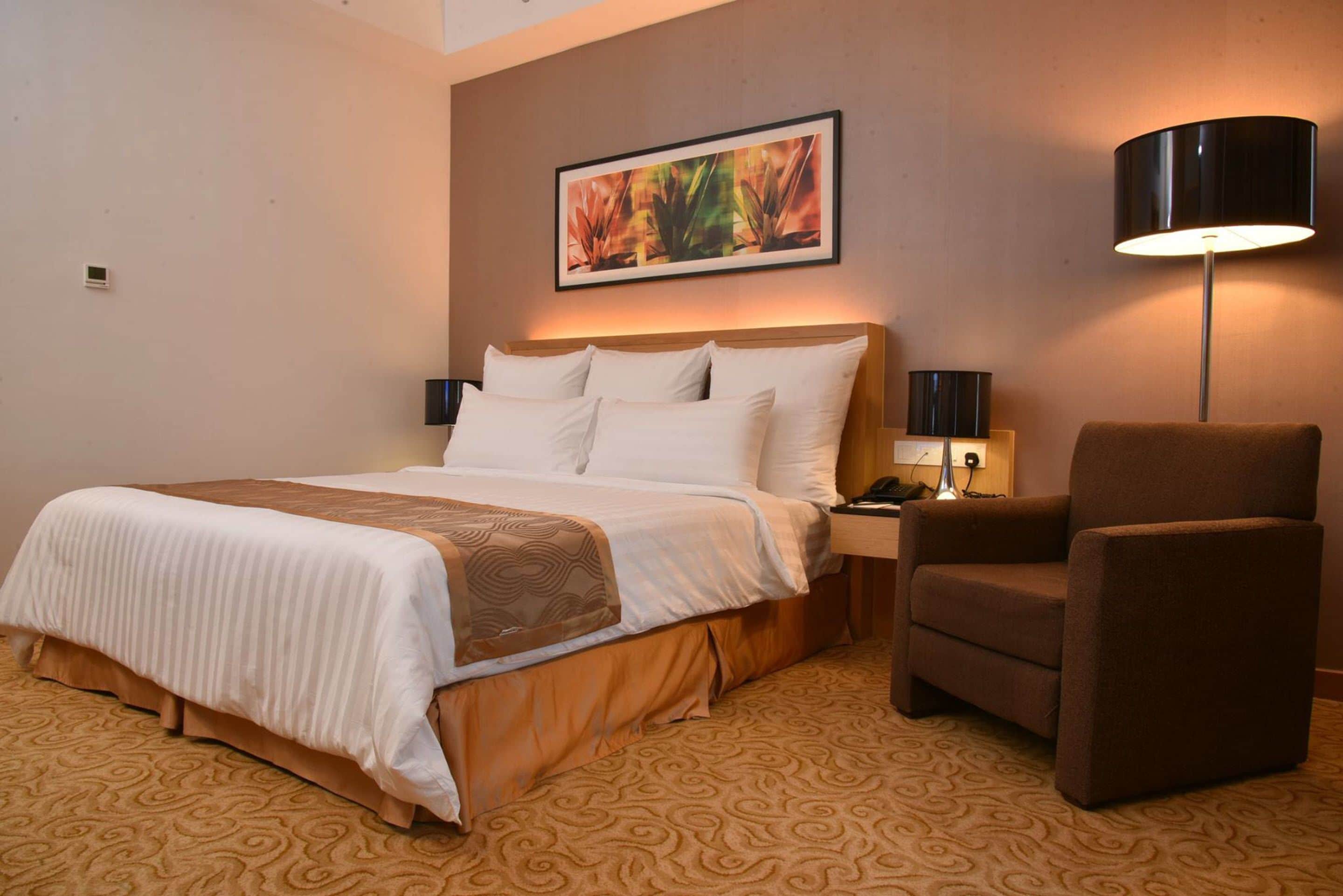 A hotel room with a bed, a chair, a nightstand, a lamp, and a painting on the wall.