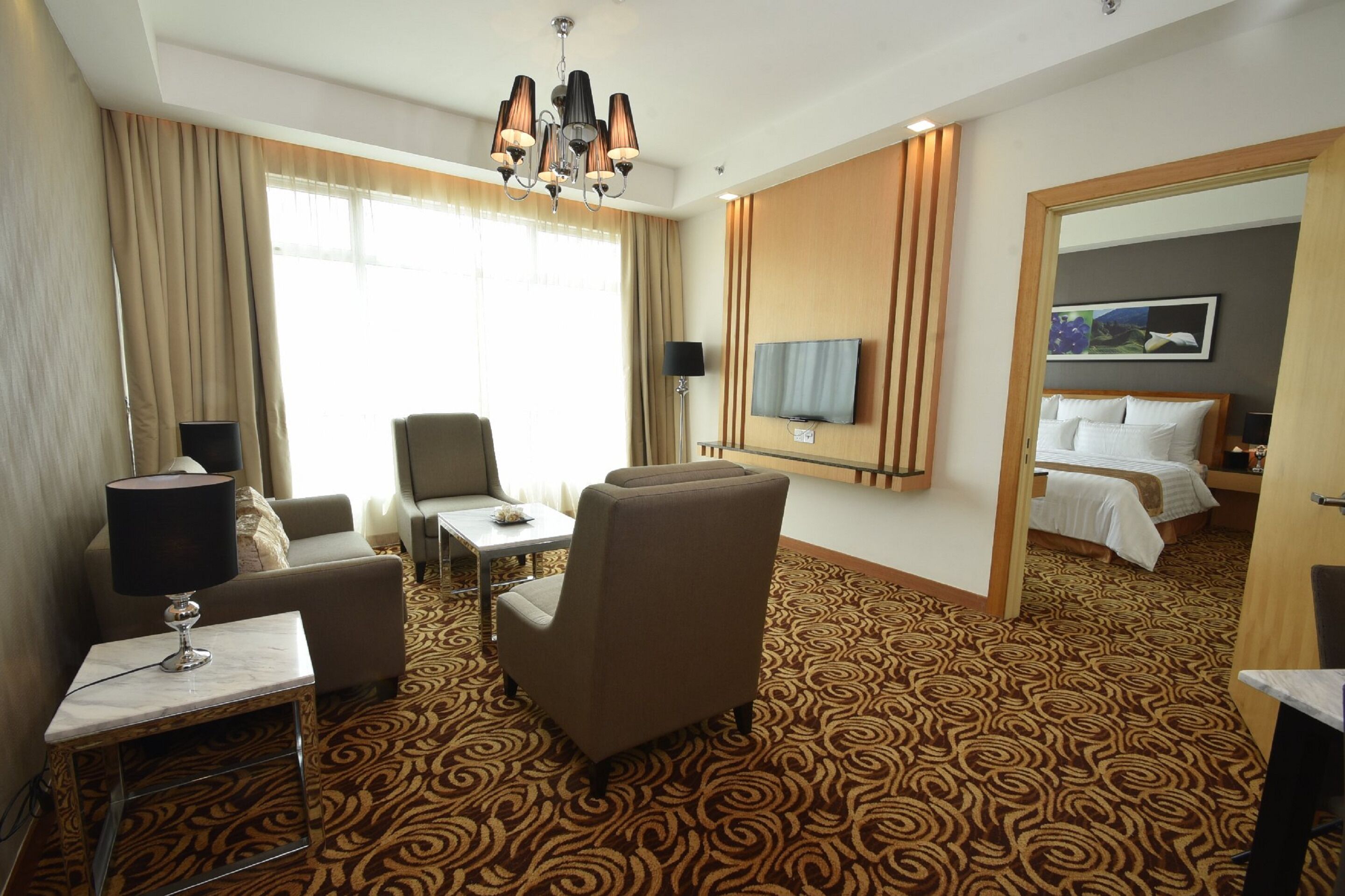 A hotel room with a patterned carpet, a sofa, a chair, a small table with a lamp, a TV, and a bed with a headboard.