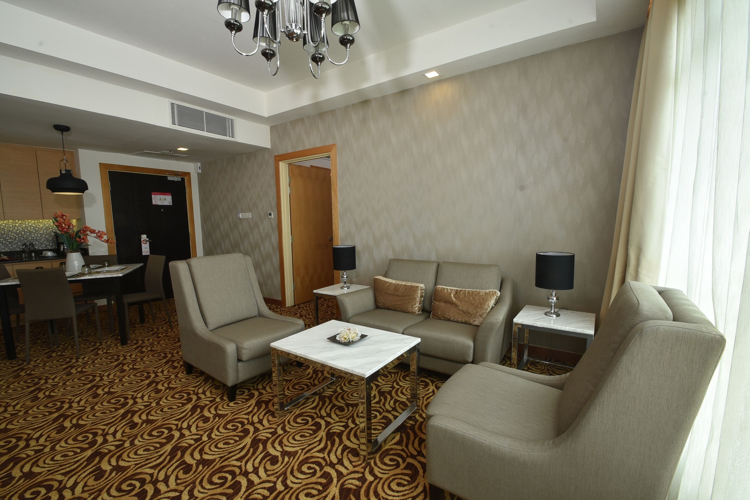 A hotel room with a patterned carpet, a sofa set, a small table, a dining area, and a chandelier.