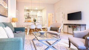 Una sala moderna con sofá, mesa de centro y sillas, una zona de comedor con mesa y sillas, una cocina al fondo y un televisor montado en la pared.